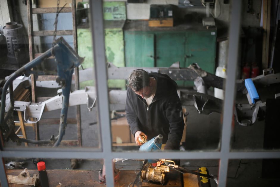 From window view of adult male worker in workwear standing at workbench and fixing metal detail while working in modern garage