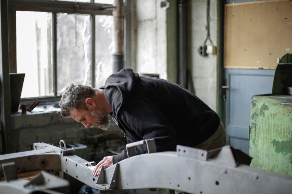 Side view of serious adult bearded mechanic in workwear standing near metal construction and working with details in spacious garage